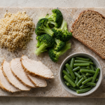 Stone cutting board with cooked brown rice, fresh broccoli florets, whole grain bread, sliced roasted turkey breast, and a bowl of green beans
