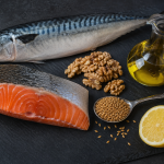 Slate cutting board with fresh salmon fillet, whole mackerel, walnuts, flaxseeds, half a lemon, and a glass cruet of golden oil