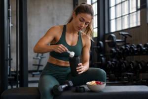 Woman preparing a creatine drink in a shaker bottle beside dumbbells and a healthy snack on a gym bench