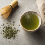 Ceramic cup of steaming green tea on a stone surface with loose green tea leaves and a bamboo whisk beside it