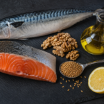 Slate cutting board with fresh salmon fillet, whole mackerel, walnuts, flaxseeds, half a lemon, and a glass cruet of golden oil