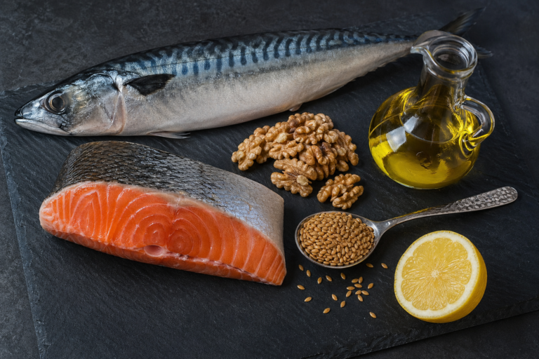 Slate cutting board with fresh salmon fillet, whole mackerel, walnuts, flaxseeds, half a lemon, and a glass cruet of golden oil