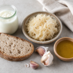 Overhead still life of kefir in a glass jar, sauerkraut, sourdough bread, garlic cloves, and olive oil on a light stone surface